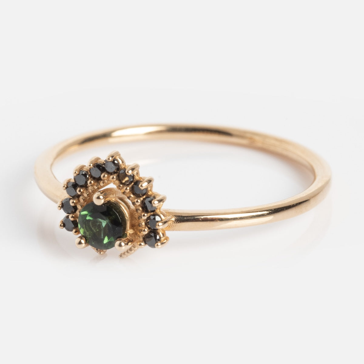 The Tosia Tourmaline Ring is a delicate solid gold ring featuring a central dark green tourmaline and a curved halo of small black diamonds, displayed on a white background.