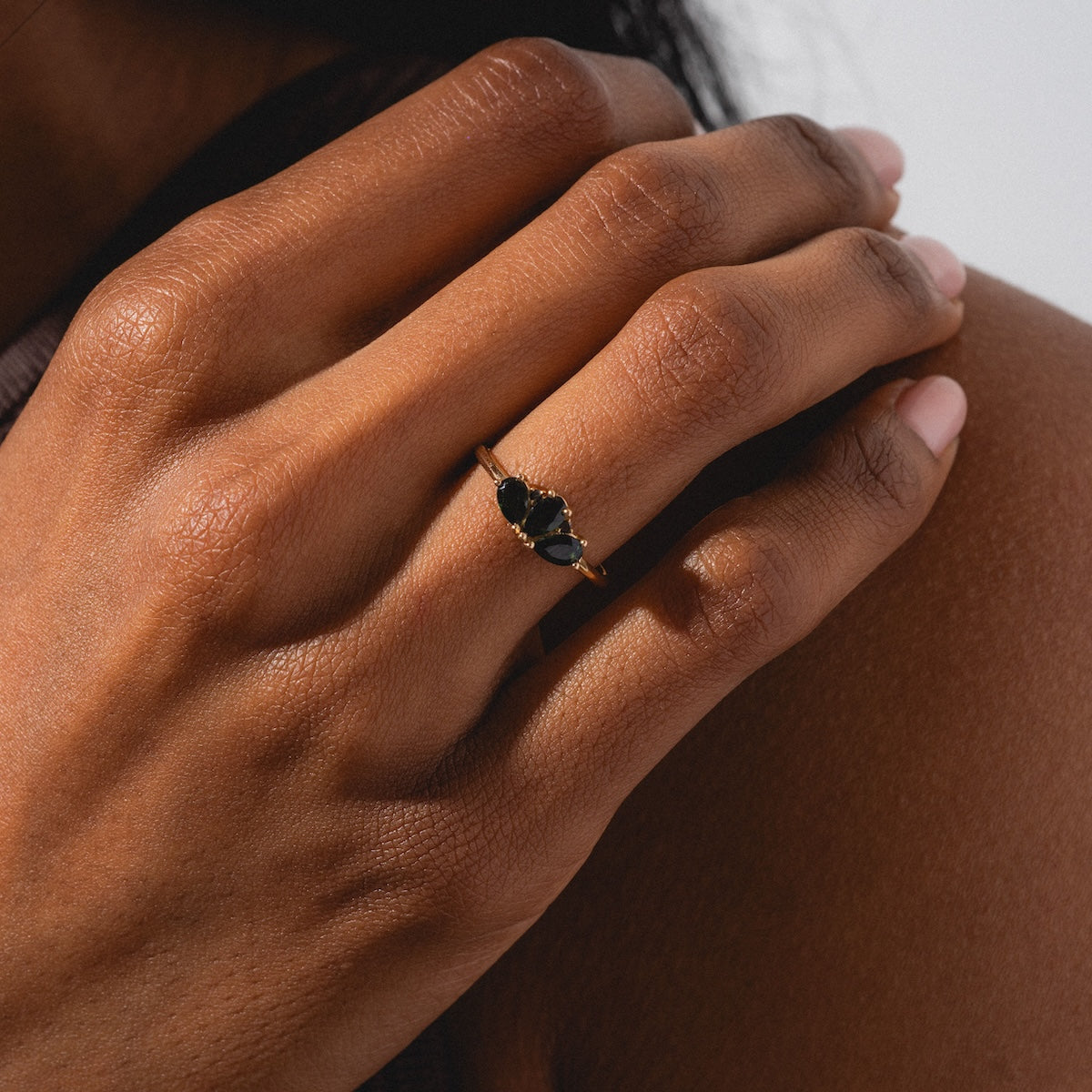 A close-up shows a hand wearing the Ava Black Diamond Ring in 14k solid gold, featuring three black stones and resting on a shoulder—an elegant choice for lovers of black diamond rings.