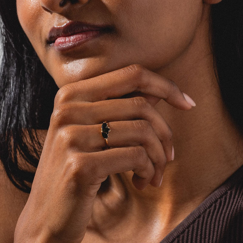 A woman with dark hair rests her chin on her hand, displaying the Ava Black Diamond Ring—a 14k solid gold ring with a dark oval gemstone. She wears a sleeveless brown top and minimal, natural makeup.