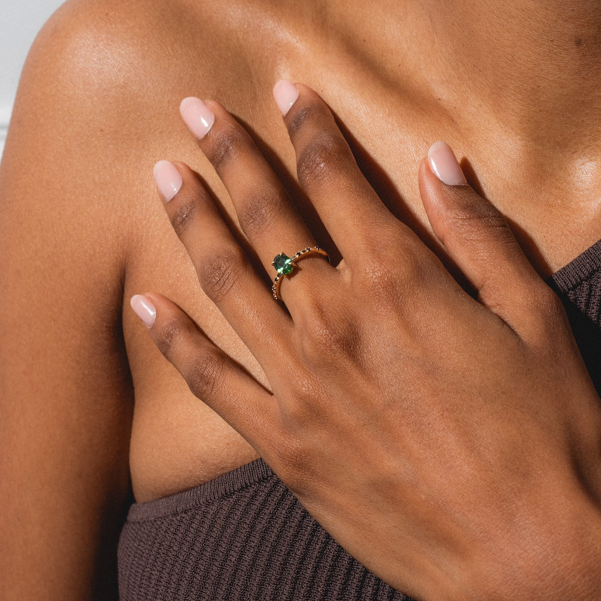 A person with light pink nail polish rests their hand on their chest, wearing the Iona Ring Black Diamond Ring in 14k yellow gold. They are dressed in a brown, textured strapless top.