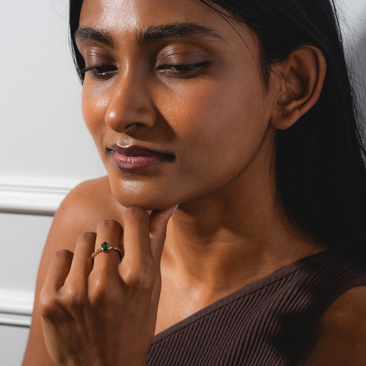 A woman with long dark hair and smooth skin rests her chin on her hand, wearing the Iona Ring Black Diamond Ring set in 14k yellow gold. She looks calm and is dressed in a one-shoulder brown top.