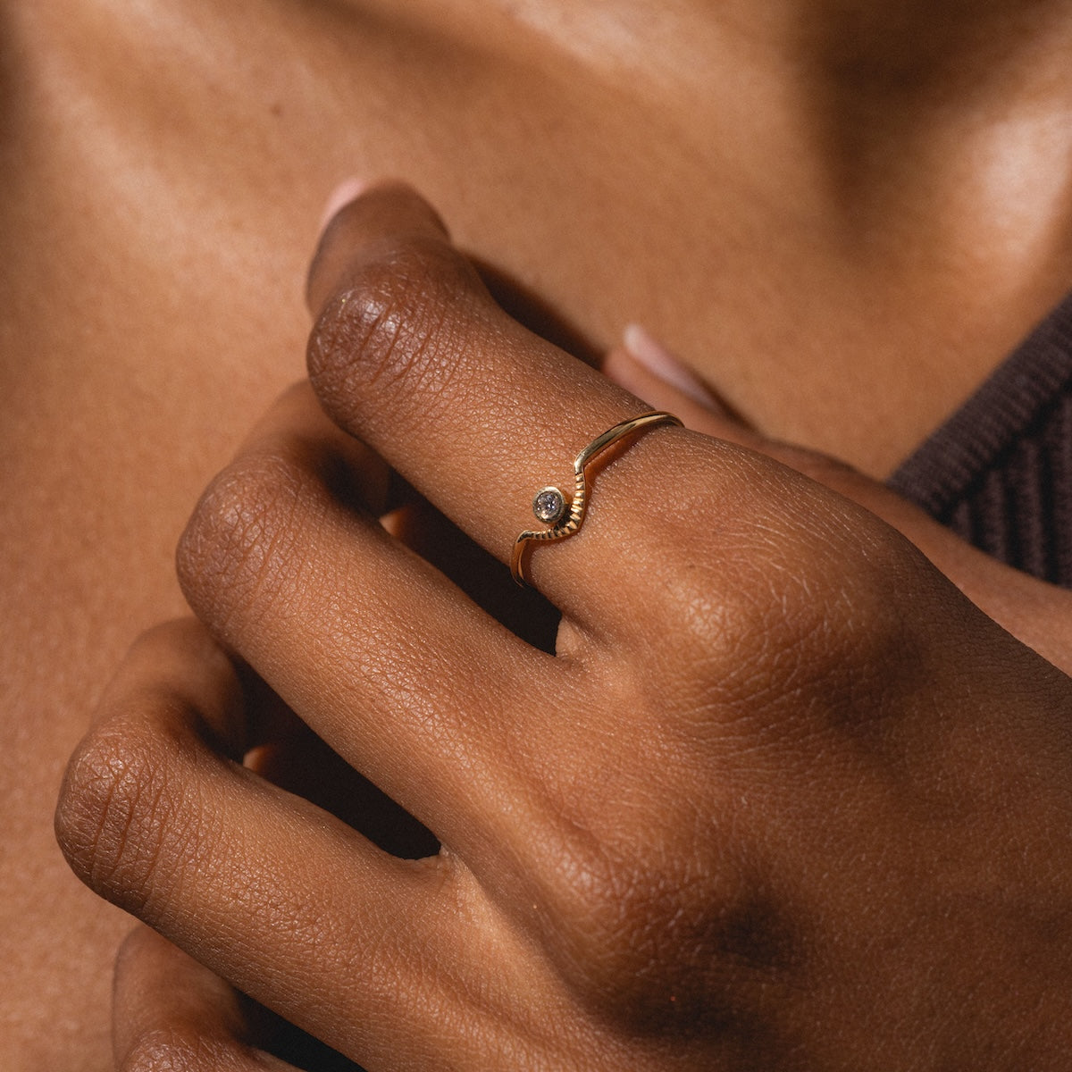 A close-up of a hand wearing the Corrie Ring, a delicate gold band with a small gemstone, highlights the jewelry against brown skin and a softly blurred dark background.