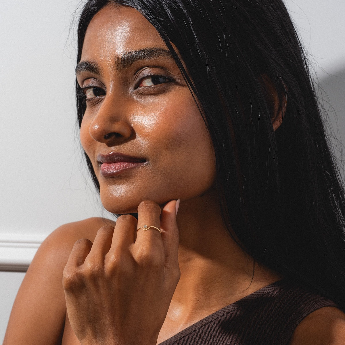 A woman with long black hair and glowing skin smiles softly at the camera. She wears a brown one-shoulder top and rests her hand, adorned with the Corrie Ring, on her chin against a light background.