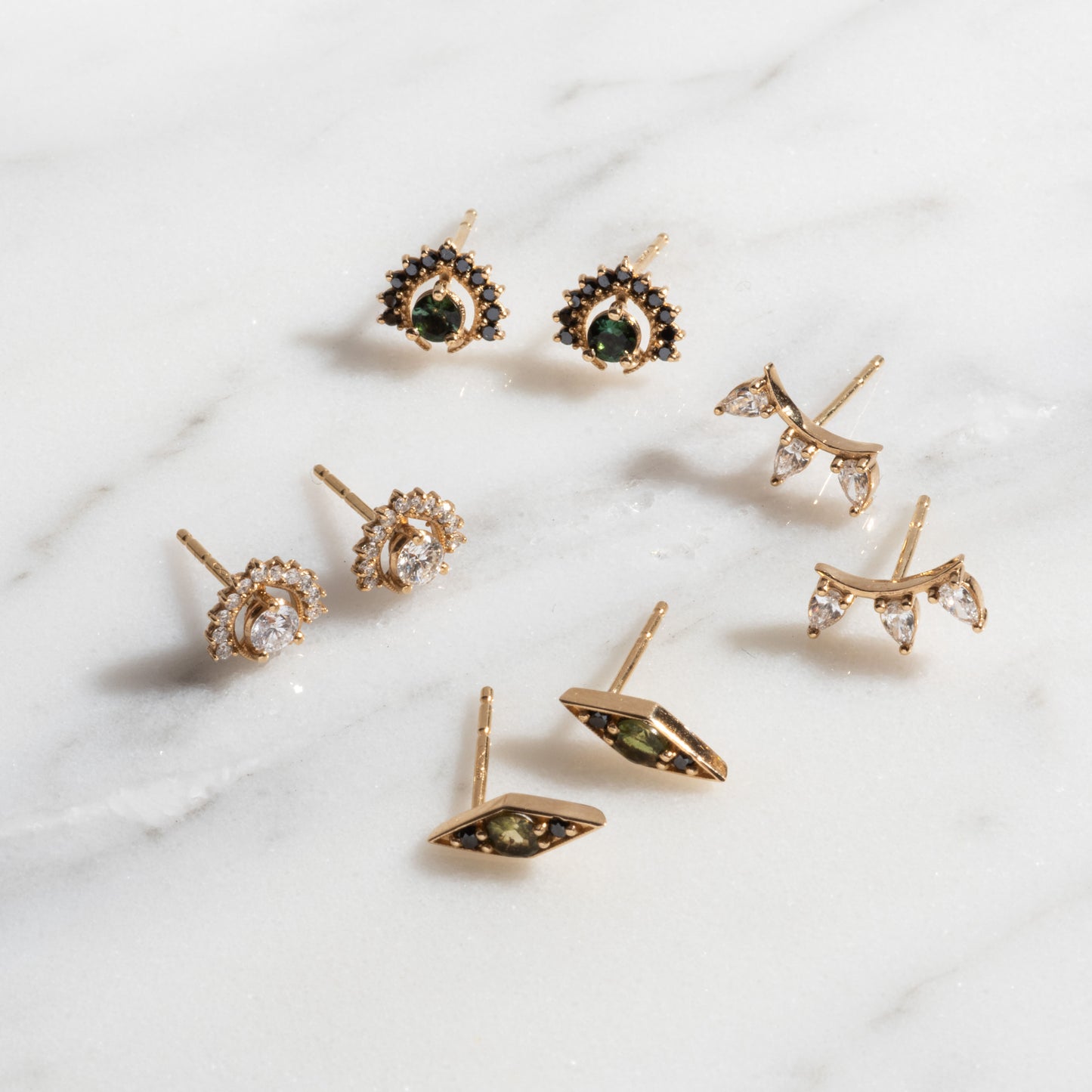 Four pairs of Tosia Diamond Studs in gold, featuring green, black, and clear gem designs, are displayed on a white marble surface.