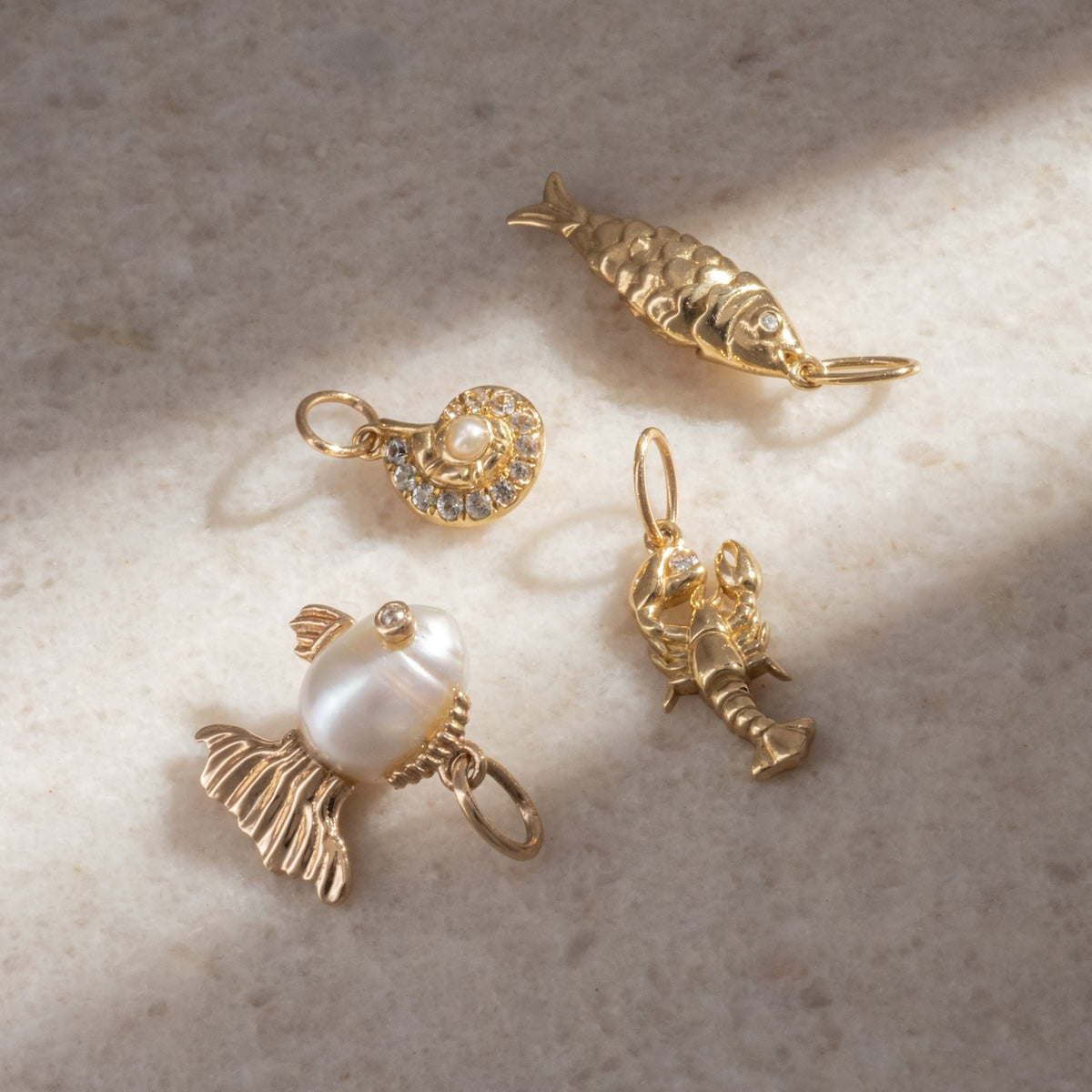 Four Solid Gold Nature Charms are displayed on a light stone surface: a fish, a hat with a pearl, a pearl fish with gold fins, and a lobster—distinct pieces for lovers of spiritual jewelry.