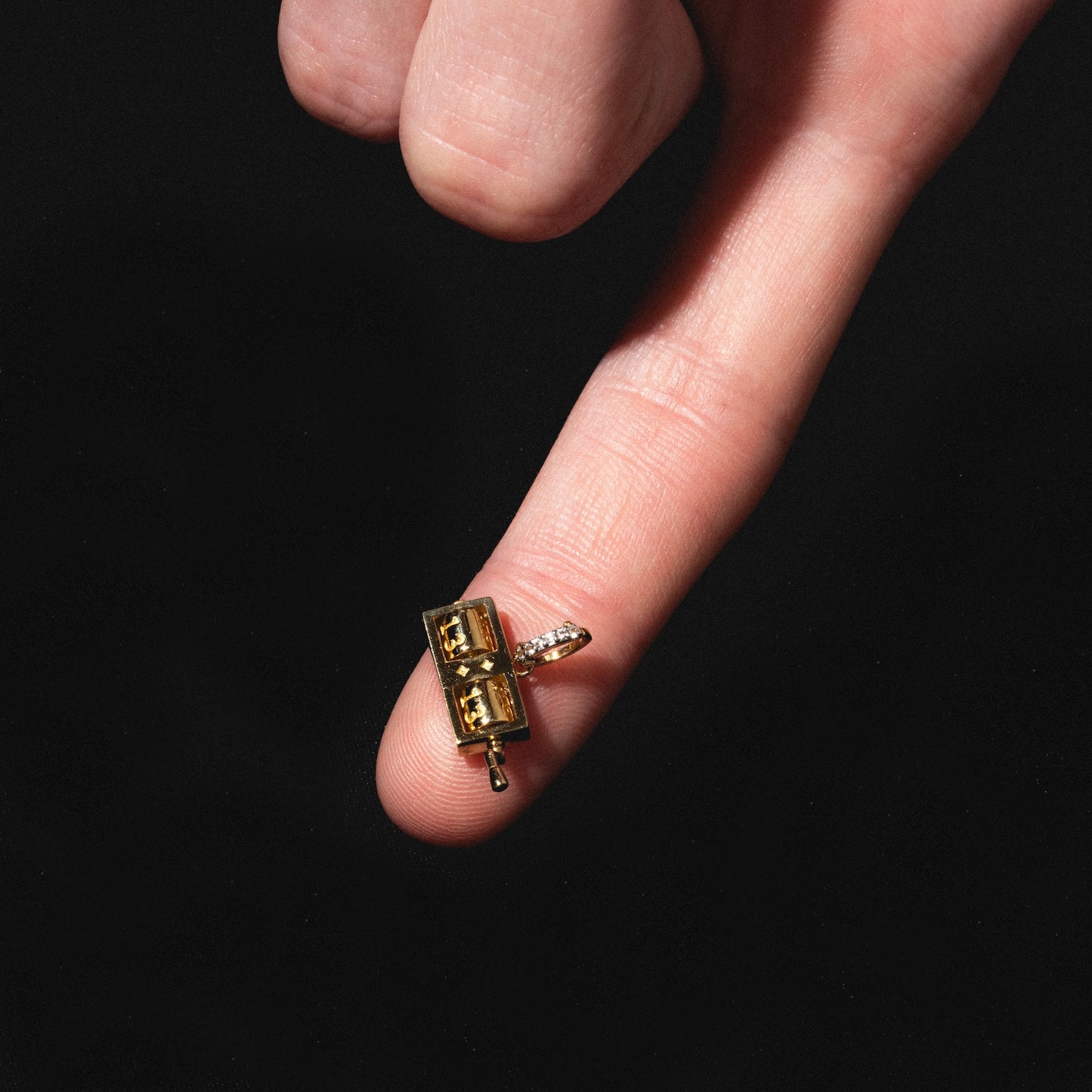 A hand with an extended index finger balances the Solid Gold Lucky Charm, shaped like a suitcase, against a black background.