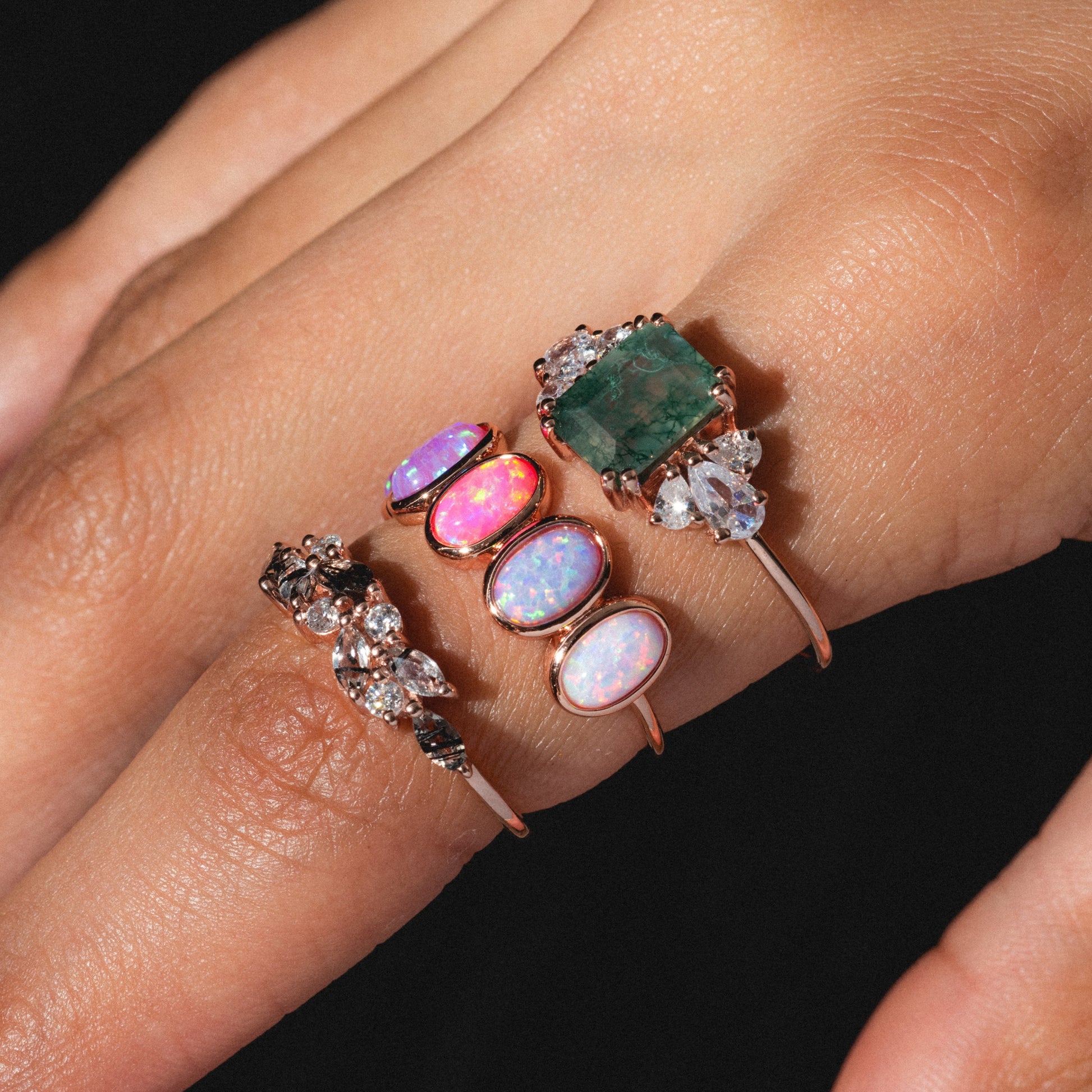 A close-up of a hand with light brown skin wearing four Gemstone Orchard Rings—gold vermeil bands set with colorful gemstones like opals and an emerald-green stone—against a black background. Nails are painted pale blue.