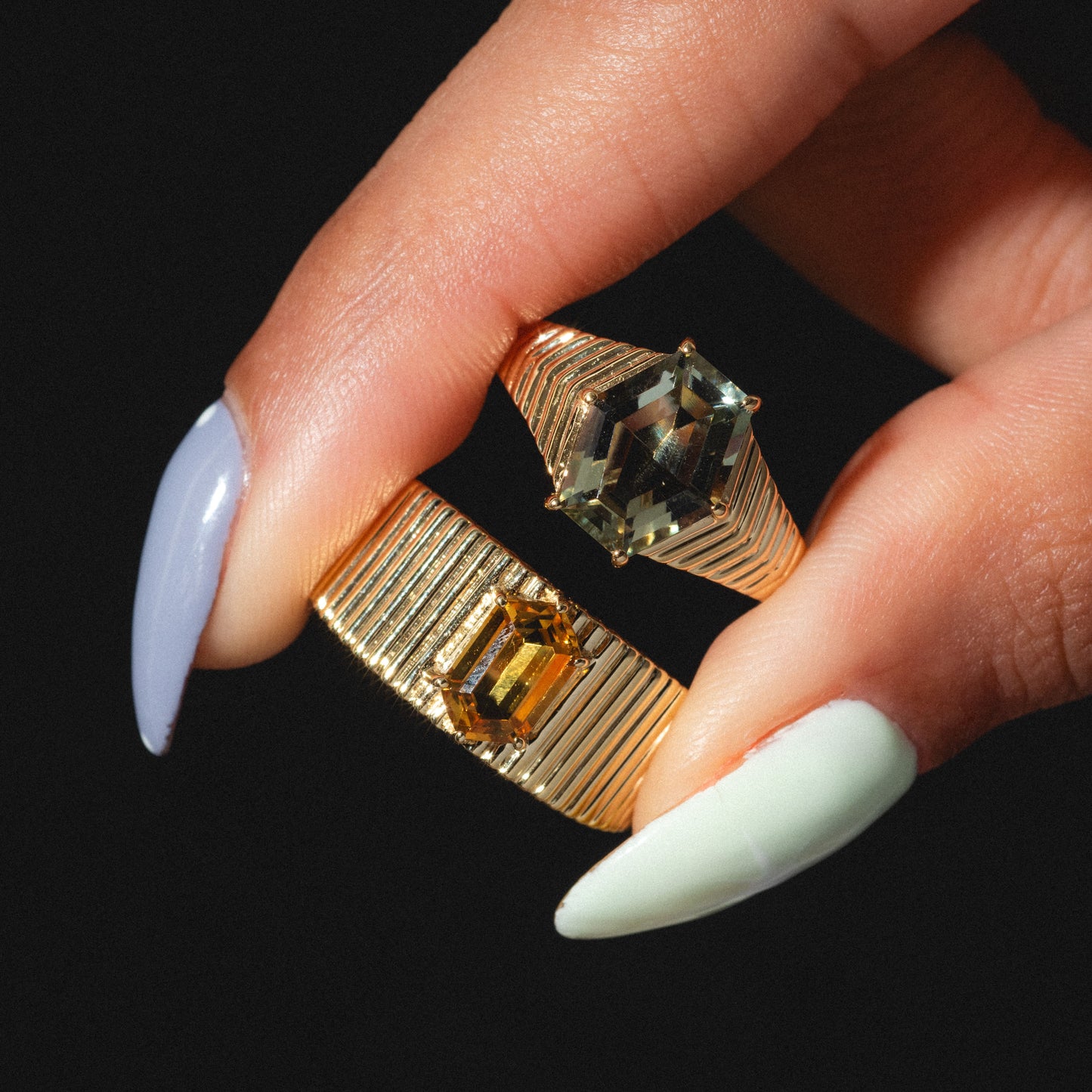 A hand with pastel-colored nails holds the Solid Gold Citrine Bold Fluted Cigar Band, a 9k yellow gold ring with bold rectangular green and yellow citrine gemstones, shown against a dark background.