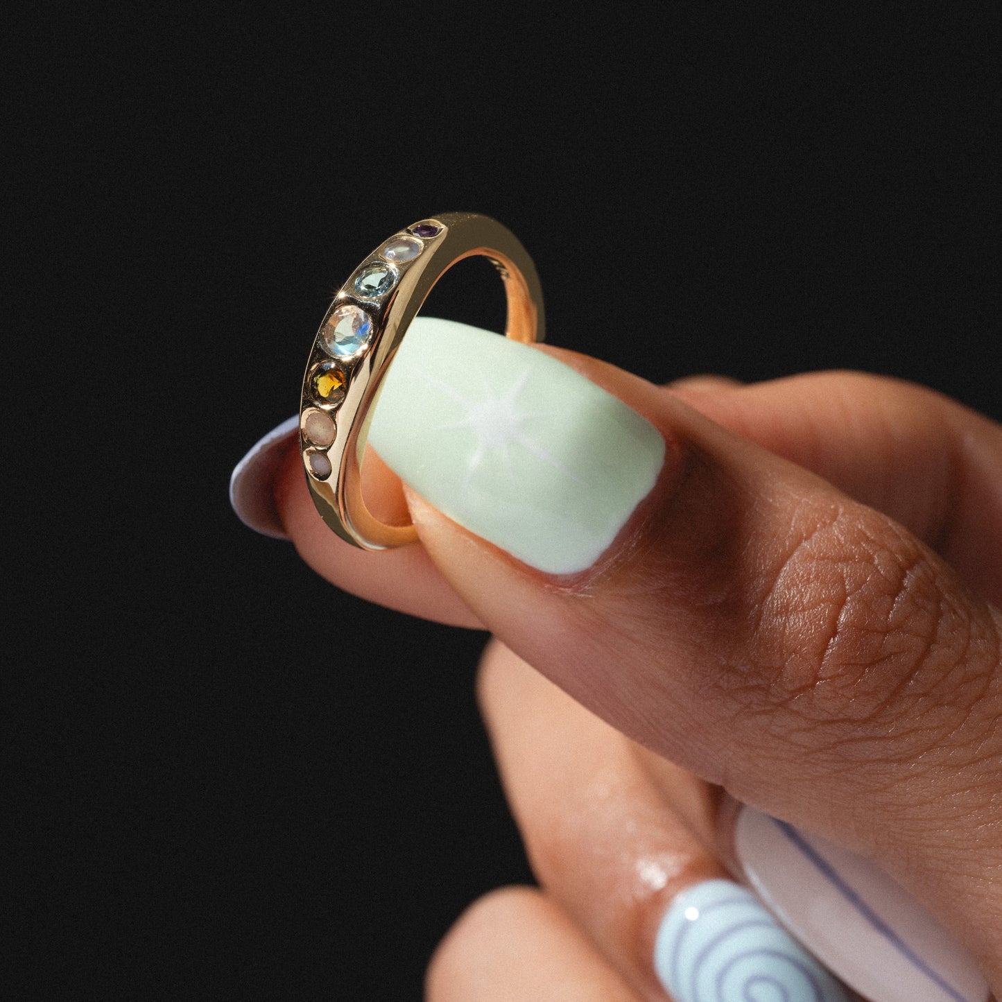 A hand with light green and white nail art holds the She's an Artist Ring in Yellow Gold, showcasing five vibrant gemstones set in solid gold, against a black background.