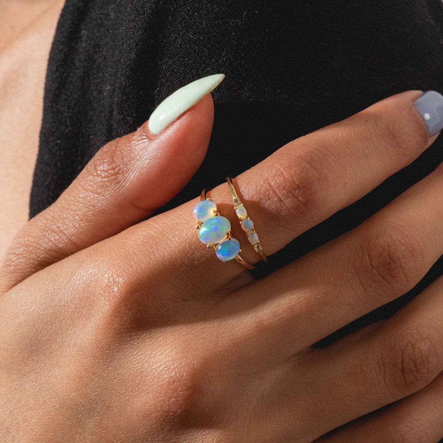 A hand with pale green and blue manicured nails showcases the Solid Gold 2025 October Capsule Opal Ring Set, featuring multiple iridescent opals, worn against a black fabric background. Comes with a free gift.
