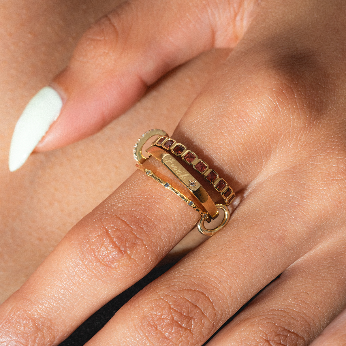 A close-up of a light-skinned hand with white nails wearing three stacked gold rings, including the Solid Gold Tiny Fuck Ring, a stacking ring with red stones, and one with small green stones.