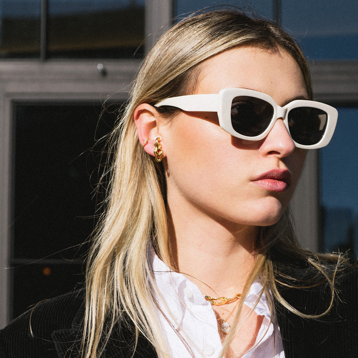 A woman with long blonde hair wears white rectangular sunglasses, a white-collared shirt with a black jacket, and the Brooklyn Earrings Sample in 14k yellow gold, standing outdoors in bright sunlight.