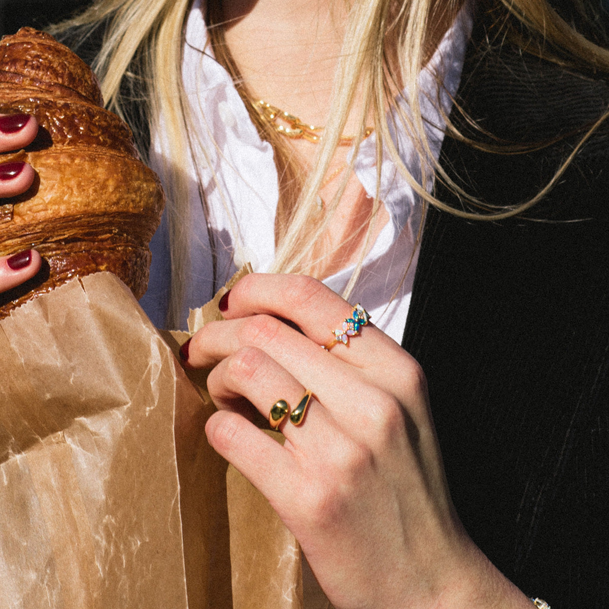 A person with blonde hair and dark polished nails, dressed in a white shirt and black jacket, wears the Opal Ombre Fleur Band and a necklace while holding a paper bag with a croissant inside.