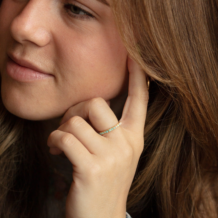 A woman with light brown hair rests her face on her hand, displaying the Indecisive Opal and Amazonite Eternity Band in yellow gold vermeil on her finger.