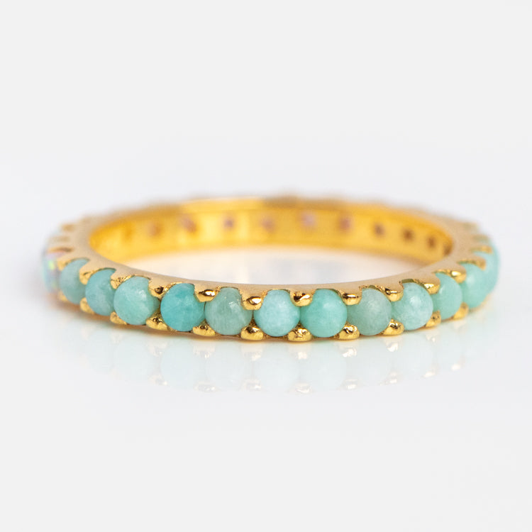 The Indecisive Opal and Amazonite Eternity Band is a yellow gold vermeil ring set with a continuous row of small, round opal and amazonite gemstones around the band, shown on a reflective white surface.