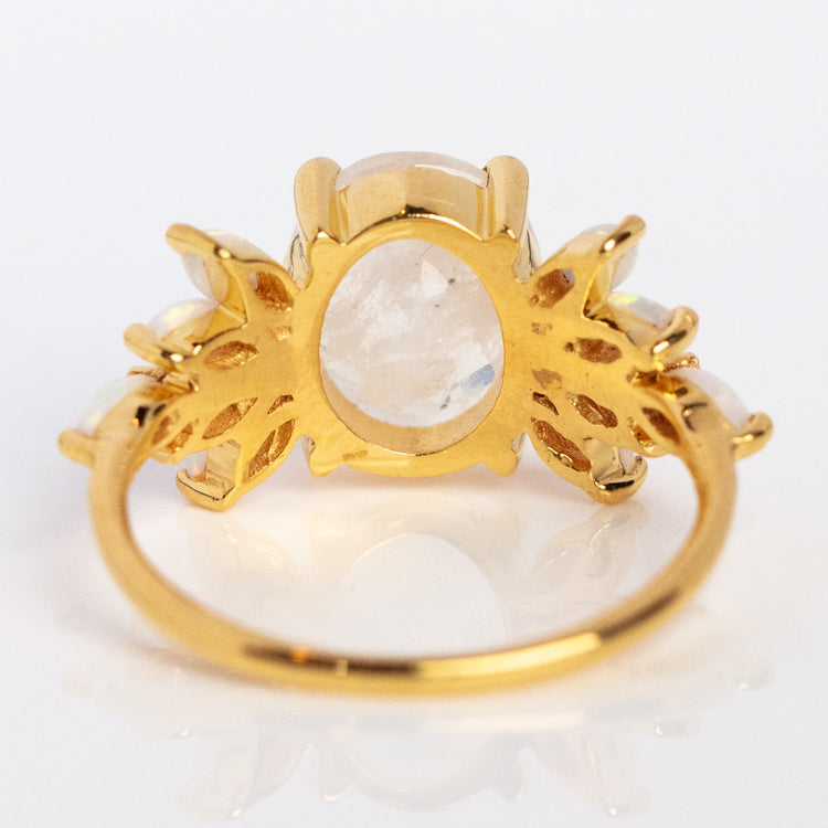 The Rainbow Moonstone and Opal Candy Crush Ring features a 14k gold vermeil band with ornate metalwork, prongs securing an oval central gemstone, and side detailing—a striking piece showcasing rainbow moonstone and opal.