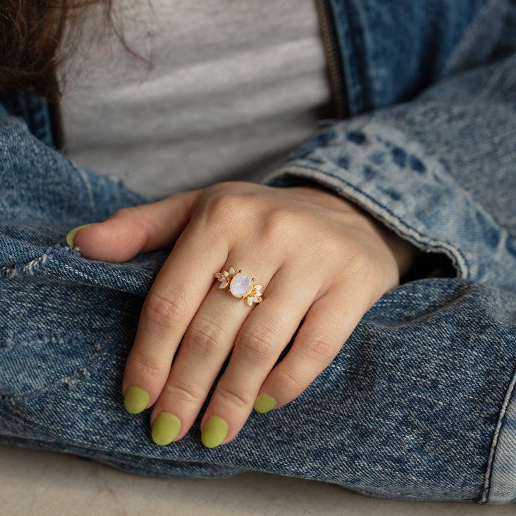 A hand with light green nails rests on a denim jacket, wearing the Rainbow Moonstone and Opal Candy Crush Ring featuring a central opal stone flanked by sparkling side gems.