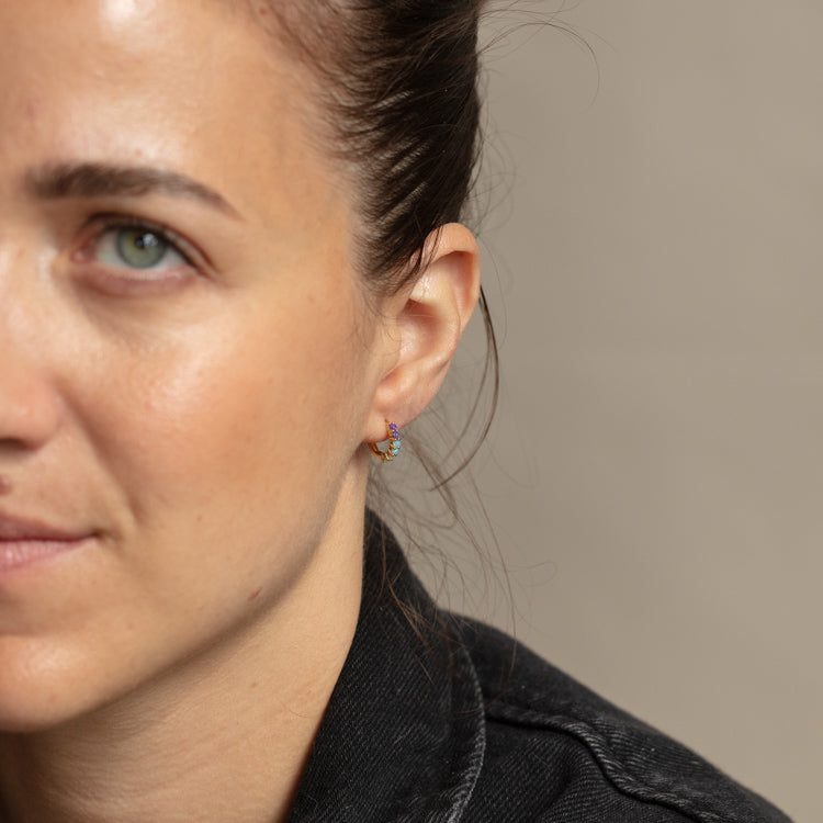 Close-up of a woman with light skin and green eyes in a dark jacket, hair pulled back to reveal the Mini Opal Ombre Huggie in 18k yellow gold vermeil. Only the left side of her face is visible.