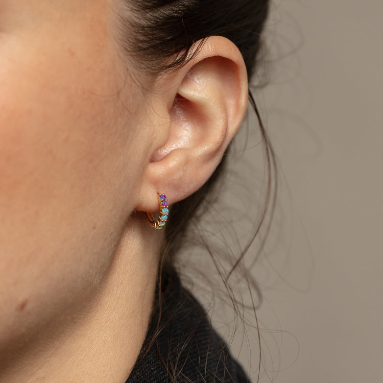 Close-up of a womans ear wearing Mini Opal Ombre Huggies in 18k yellow gold vermeil, featuring vibrant multi-colored opal gemstones. Her hair is tied back, making the earrings stand out against a neutral background.