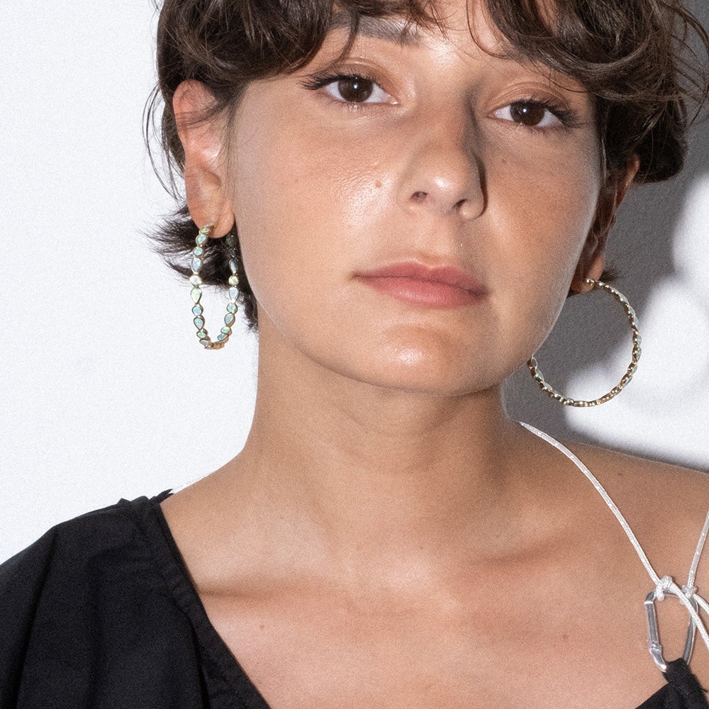 A person with short brown hair wears Isla Opal Hoop Earrings and a black top with a visible white strap, looking directly at the camera against a plain white background.