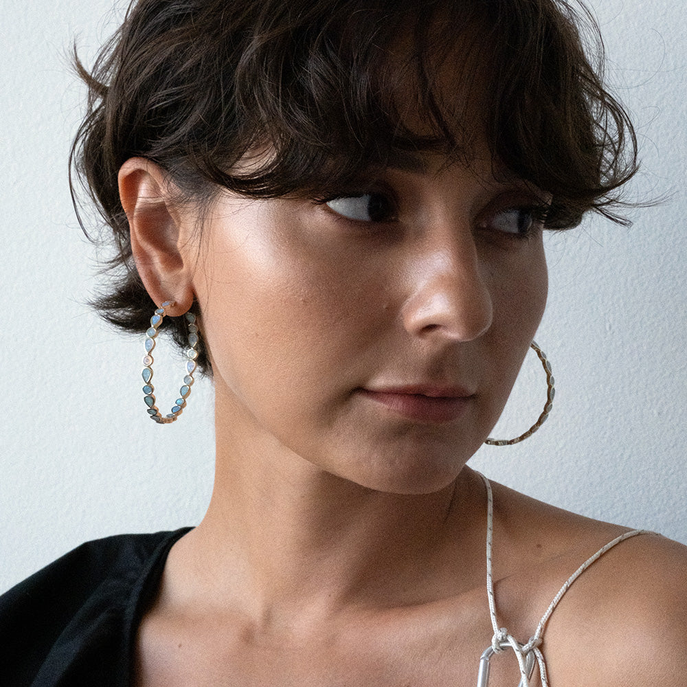 A woman with short, wavy brown hair looks to the side, wearing large Isla Opal Hoop Earrings and a strappy top against a plain, light background.