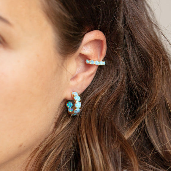 Close-up of a woman’s ear wearing two Oh She Fancy Blue Opal Huggie Hoop Earrings in 18k yellow gold—one on the lobe and a smaller one on the upper cartilage. Brown wavy hair frames the ear.