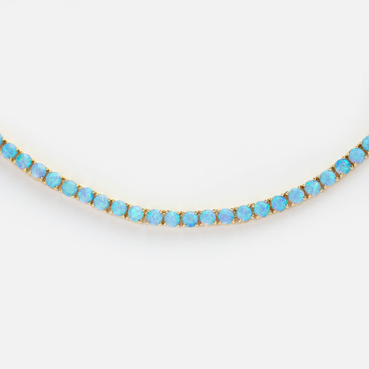 The Grand Heiress Tennis Necklace features a row of iridescent blue opals set in an elegant design with 18k gold plating, shown on a white background.