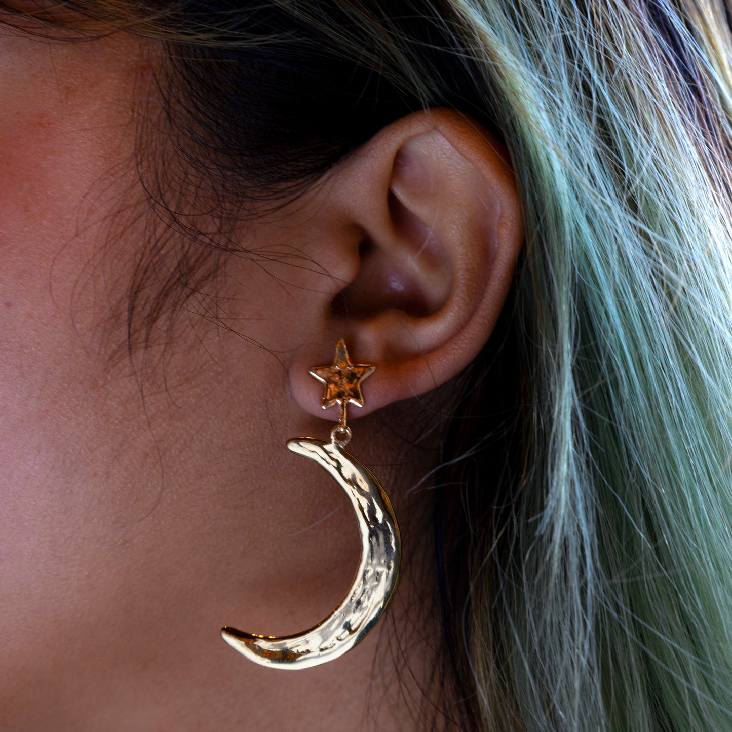 A close-up of a person with light green and blonde hair wearing the Luiny x Local Eclectic Bold Star Moon Drop Earring Sample—solid brass crescent moons hanging from gold stars, 2.4 long with secure sterling silver posts for comfort.
