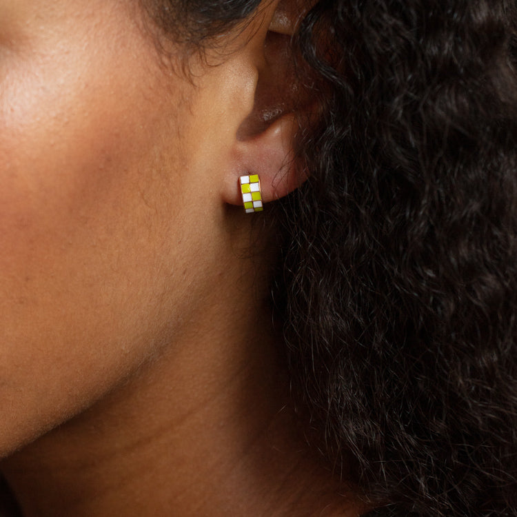 A person with curly dark hair and warm brown skin wears Checkered Huggie Earrings—small enamel hoops featuring a yellow and white checkered pattern.