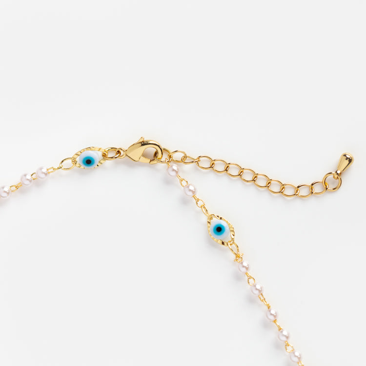 The Dainty Evil Eye Pearl Necklace features a gold-plated chain with small white beads and two blue evil eye charms, plus an adjustable clasp, shown against a white background.
