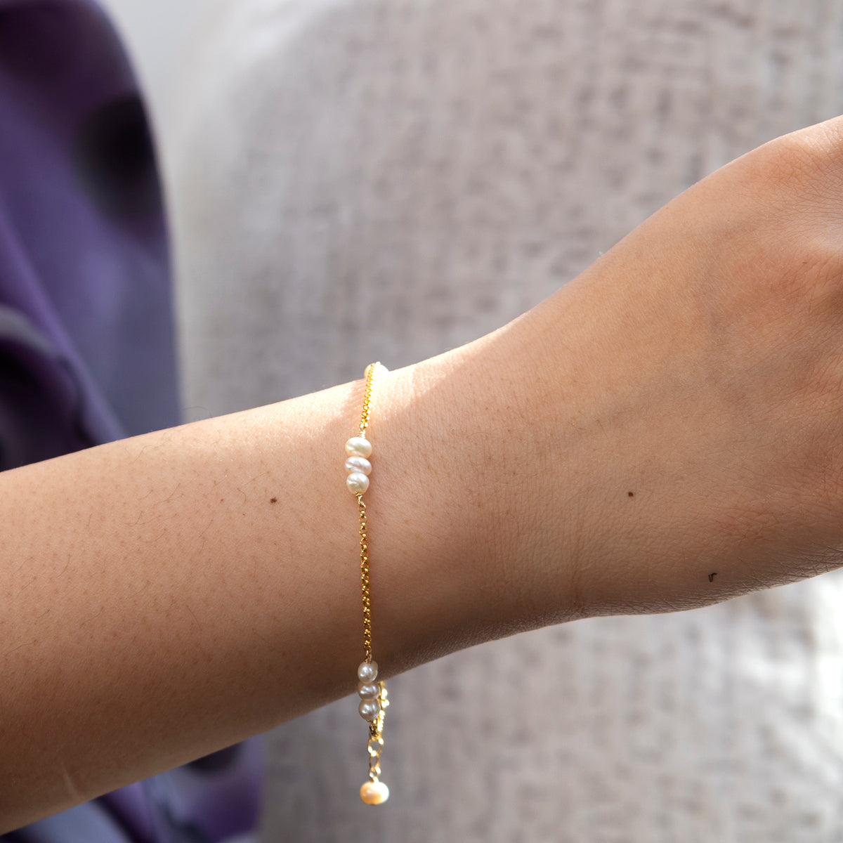 A close-up of a wrist showcases the Dottie Bracelet—crafted in 14k gold fill and adorned with small white pearls. The softly blurred background features hints of purple and light beige fabric.