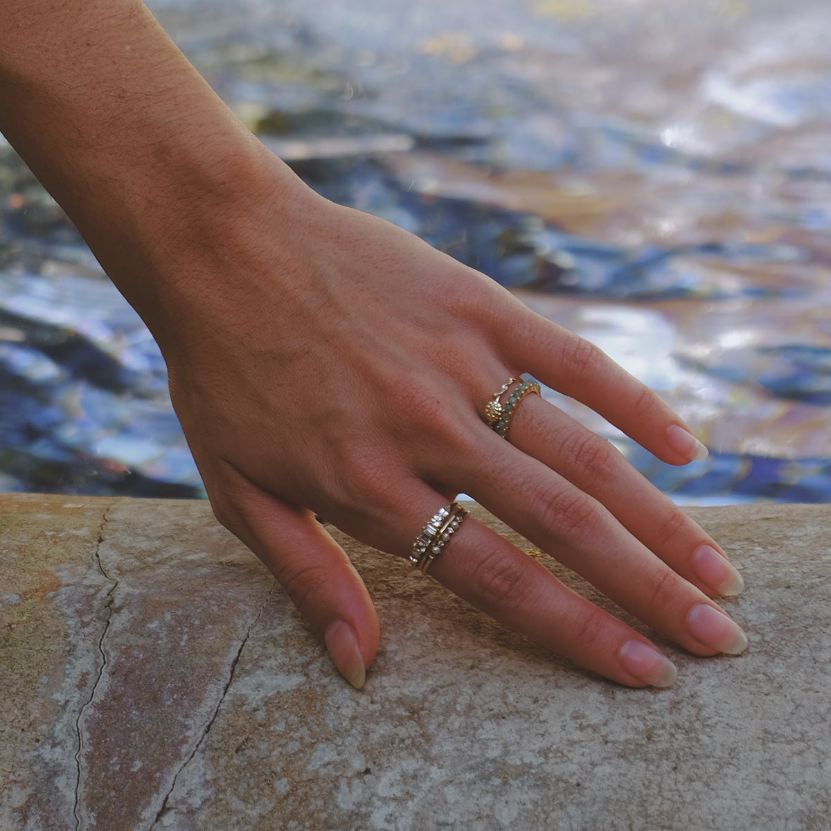 A hand with manicured nails rests on a stone surface near water, featuring the Solid Gold Birth Flower Ring and a birthstone ring on the middle and ring fingers.