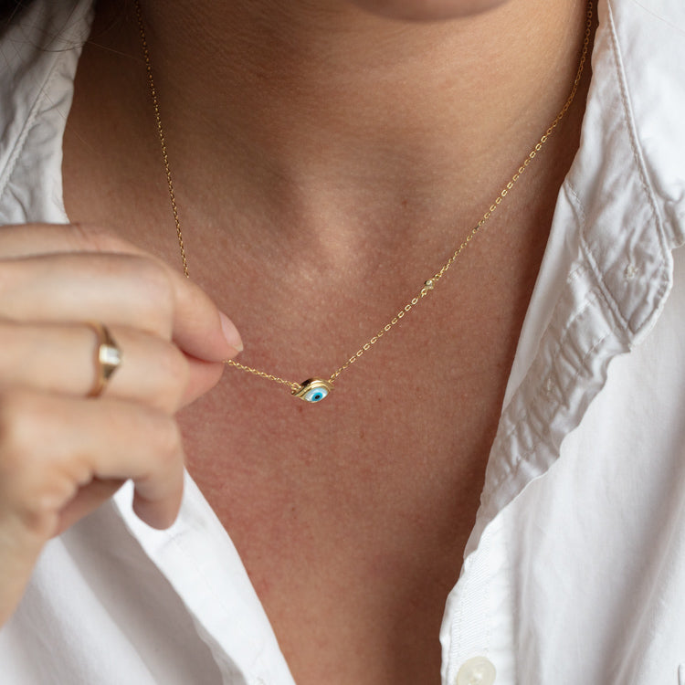 A person in a white collared shirt holds the Madison Evil Eye Necklace, a gold vermeil piece with a small pendant, close to their neck.