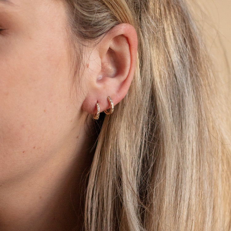 Close-up of a person’s ear with two Solid Gold Salt and Pepper Diamond Huggie Hoops Sample earrings. The hoops are 10k solid yellow gold, each encrusted with tiny salt and pepper diamonds. The model has light skin and long, blonde hair.