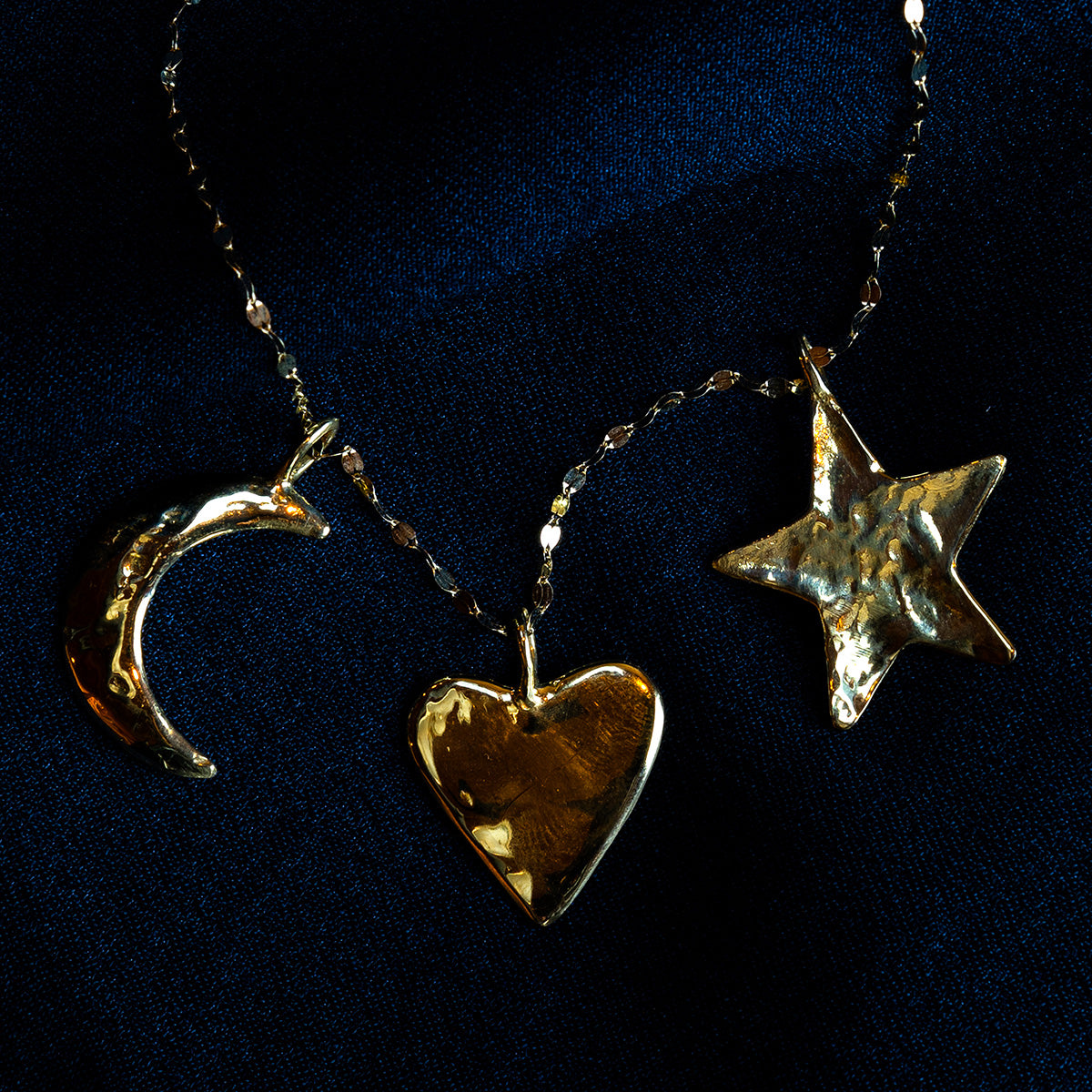 The Luiny x Local Eclectic Charms necklace with gold-tone solid brass pendants—a crescent moon, heart, and star—rests on dark blue fabric, creating a striking celestial look.