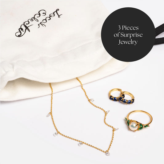 A white jewelry pouch with Secret Entity text sits beside a gold necklace with small gems and two gold rings with blue and green stones. A black circle reads: 3 Surprise Box! jewelry pieces from our exclusive collection.