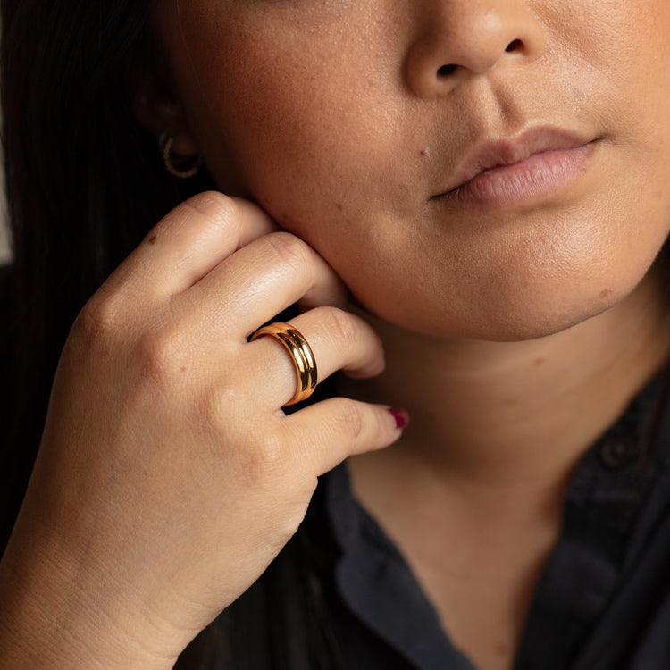 A close-up of a woman with a neutral expression, wearing the Marilou Cigar Band Ring on her index finger and a gold hoop earring, dressed in a dark top.