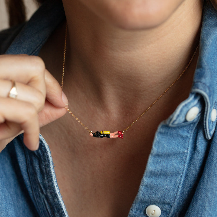 A person in a blue denim shirt holds the Scuba Diver Necklace, which features a yellow gold plated, enameled pendant shaped like a diver in a black wetsuit with a yellow tank and red fins.