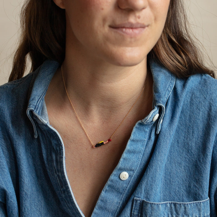 A woman wears a blue denim shirt with an open collar and the Scuba Diver Necklace—a yellow gold plated chain featuring a small, colorful enameled pendant. Her face is partly visible with a subtle smile.