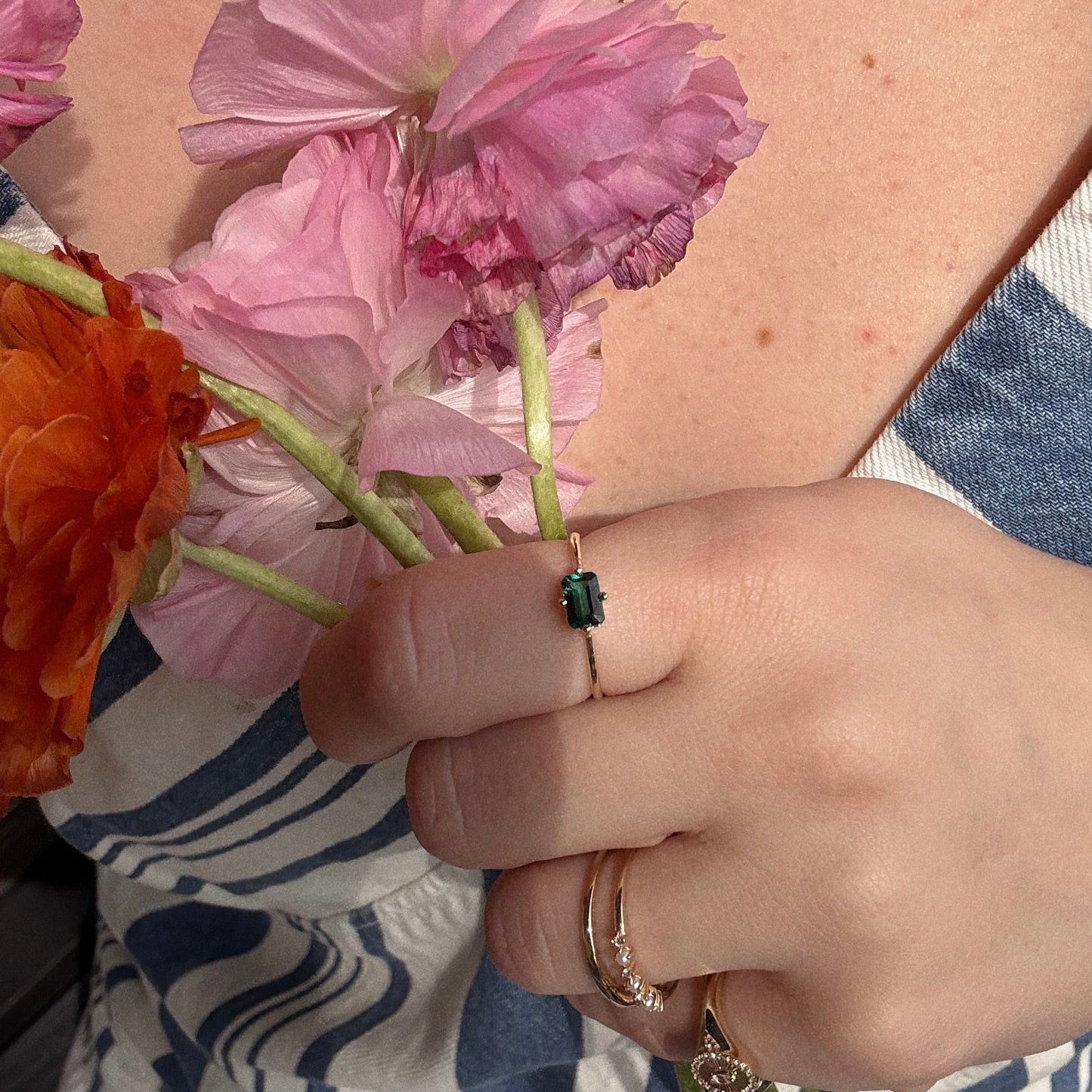 A hand holding colorful flowers, wearing the 14k Mod Love Birthstone Ring featuring a green rectangular gemstone, paired with a blue and white striped outfit.