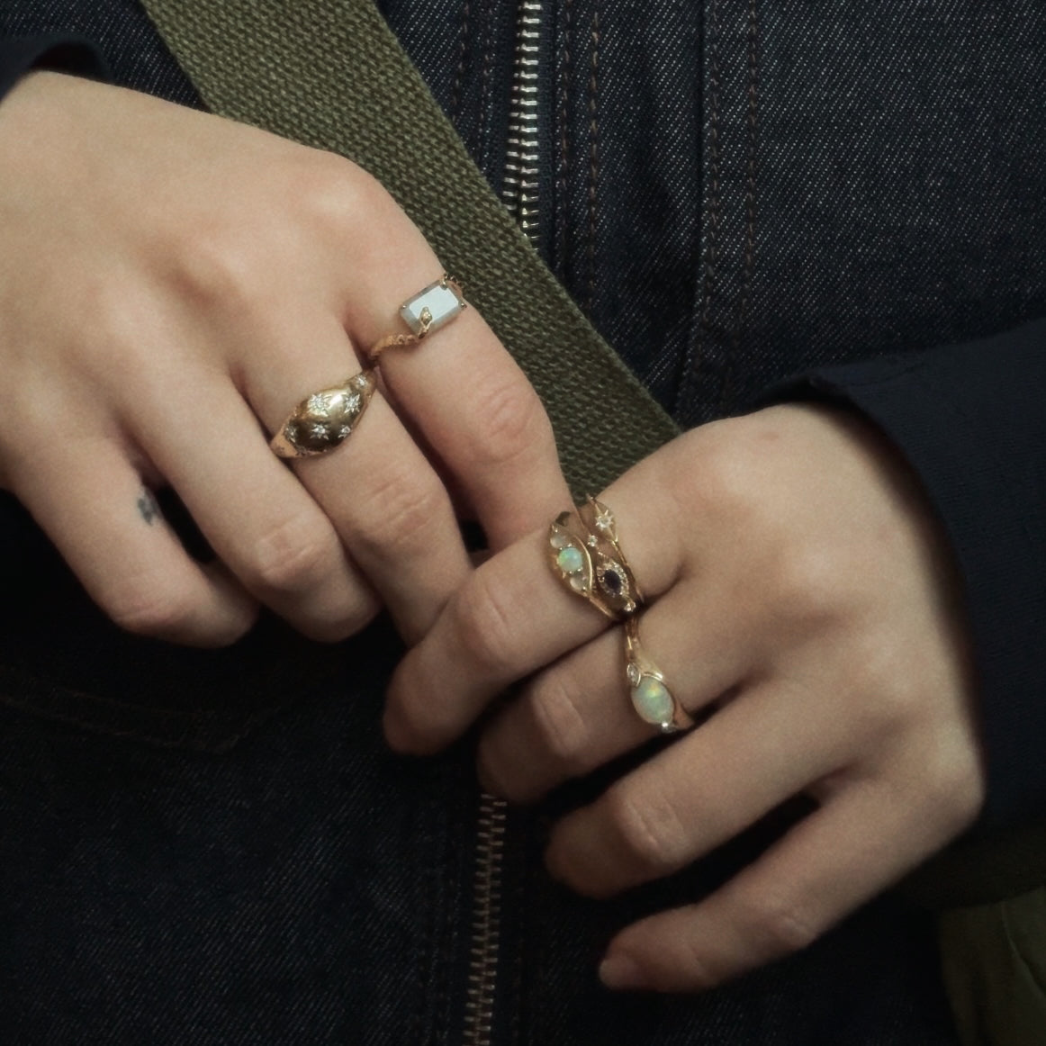 A person wears multiple ornate gold rings, including the Solid Gold Iolite Evil Eye Ring, on both hands, dressed in a dark denim jacket and holding a green strap.
