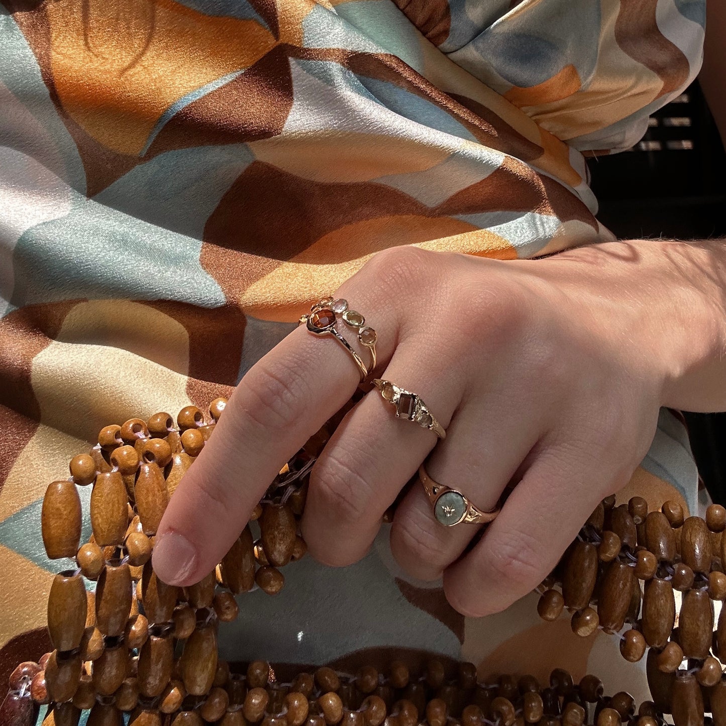 A hand adorned with the Solid Gold Bold Organic Dawn Band and two other gold rings, including a Local Eclectic exclusive, rests on a wooden beaded bag. The person wears a silky patterned dress in brown, blue, and cream tones.
