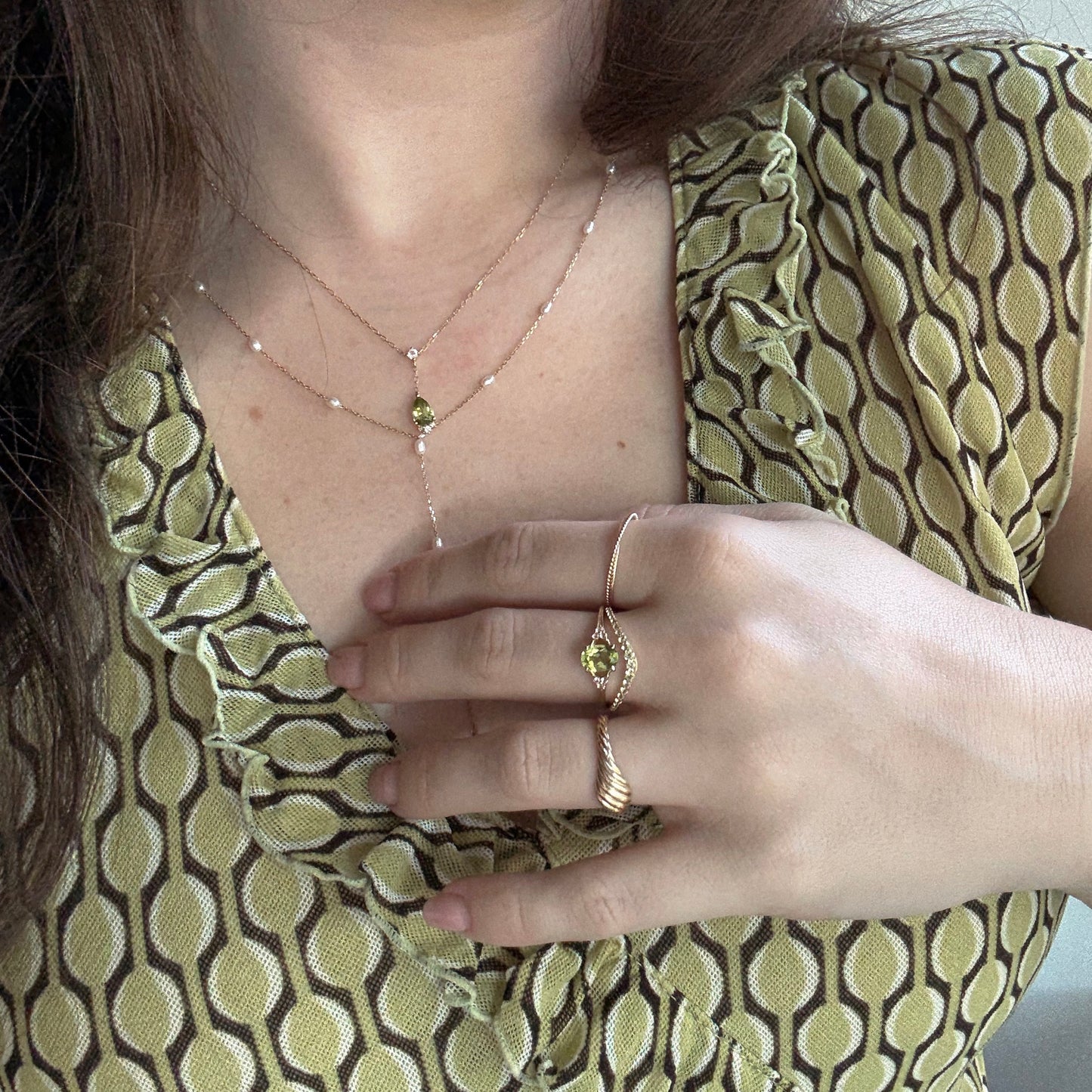 A woman in a green patterned dress showcases two rings and the Solid Gold 2025 August Capsule Vintage Inspired Peridot Drop Necklace—featuring an elegant peridot pendant that matches her ring, highlighting the August birthstone.