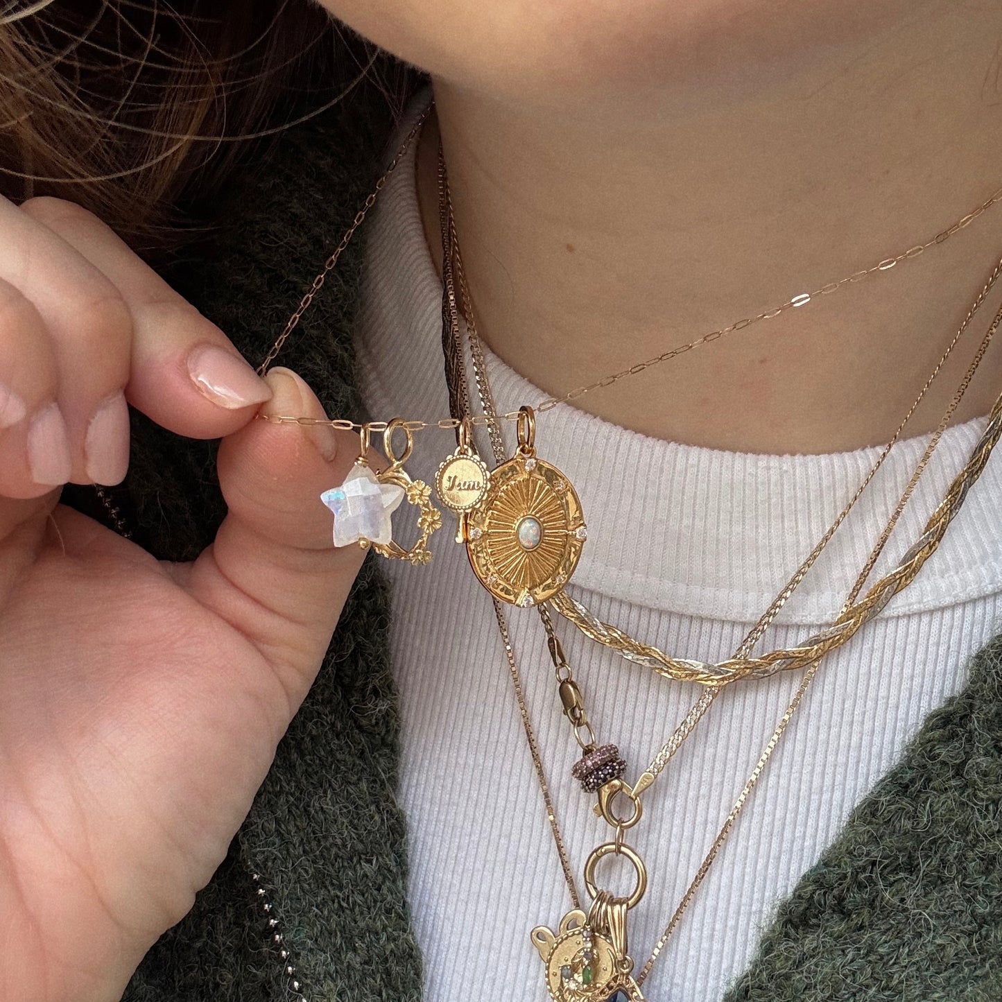 Wearing a textured green sweater, a person holds up layered gold necklaces featuring Devi Arts Collective x Local Eclectic Carved Crystal Charms, including a star, round medallion, and an engraved Mama moonstone.