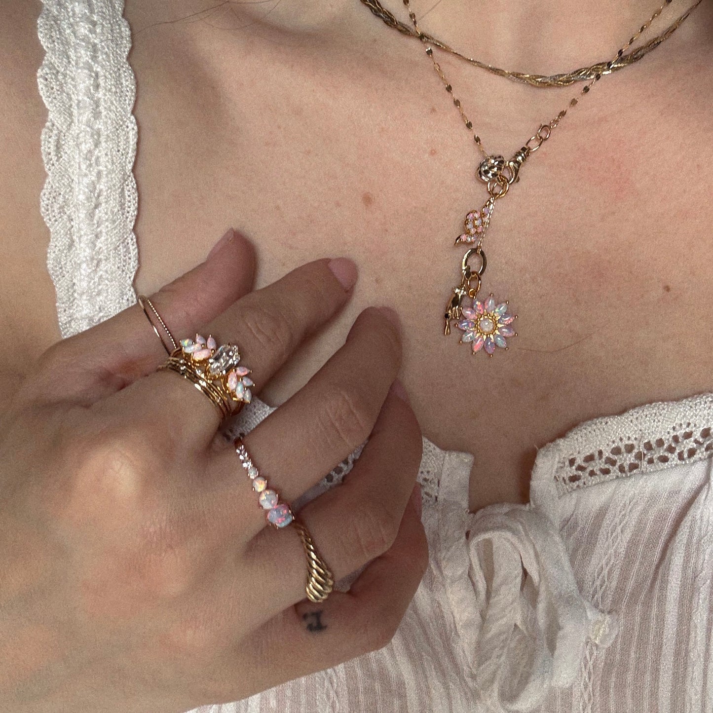 A person wearing a white textured top and layered 18k gold vermeil jewelry, including necklaces, bracelets with pink and white flower charms, and the Pink Opal Ombre and Topaz Lotus Ring. Their hand gently touches their collarbone.