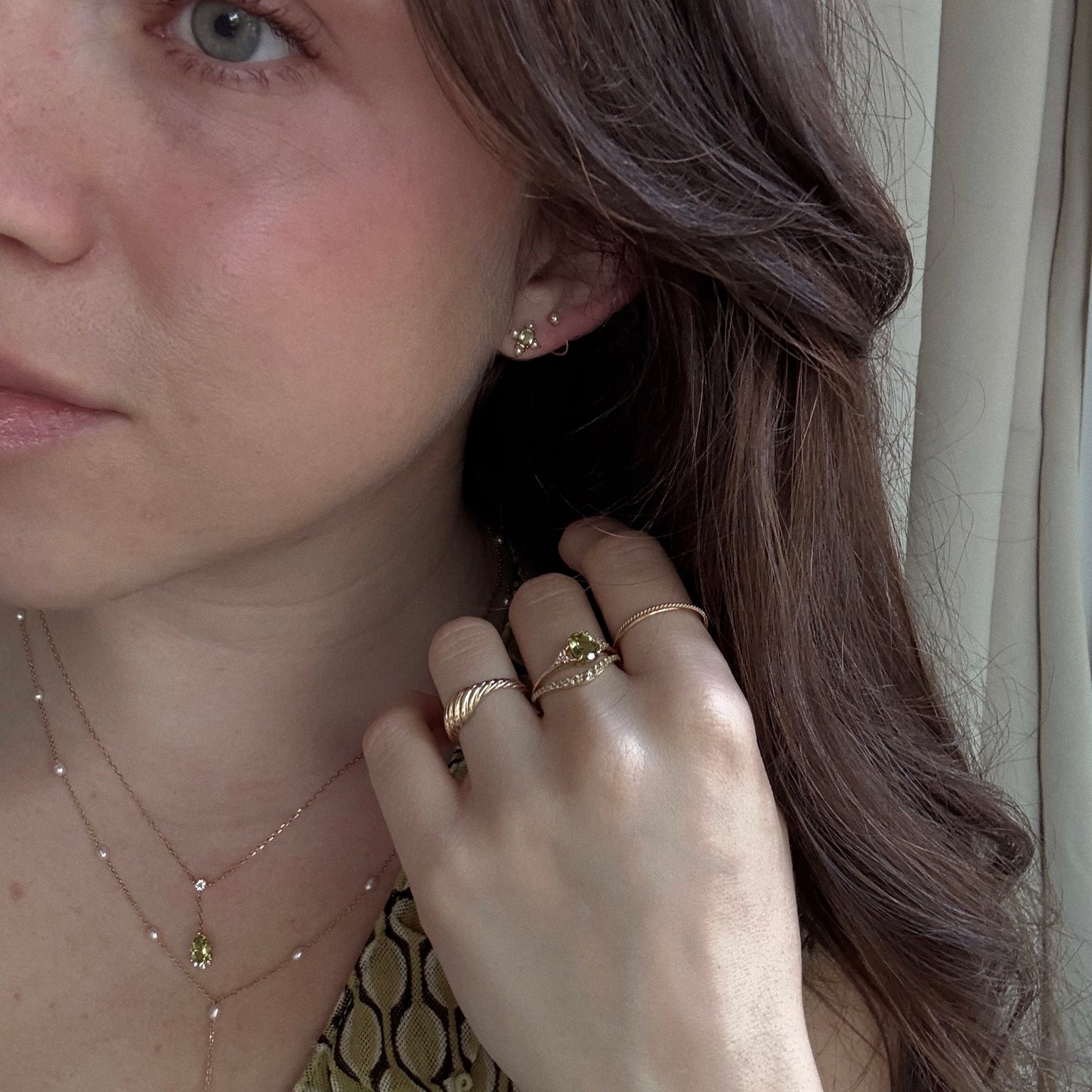A woman with long brown hair wears gold rings, necklaces, and Solid Gold 2025 August Capsule Vintage Inspired Peridot Stud Earrings. Her hand is near her face, accenting the peridot jewelry. She has light skin, a patterned top, and a curtain behind her.