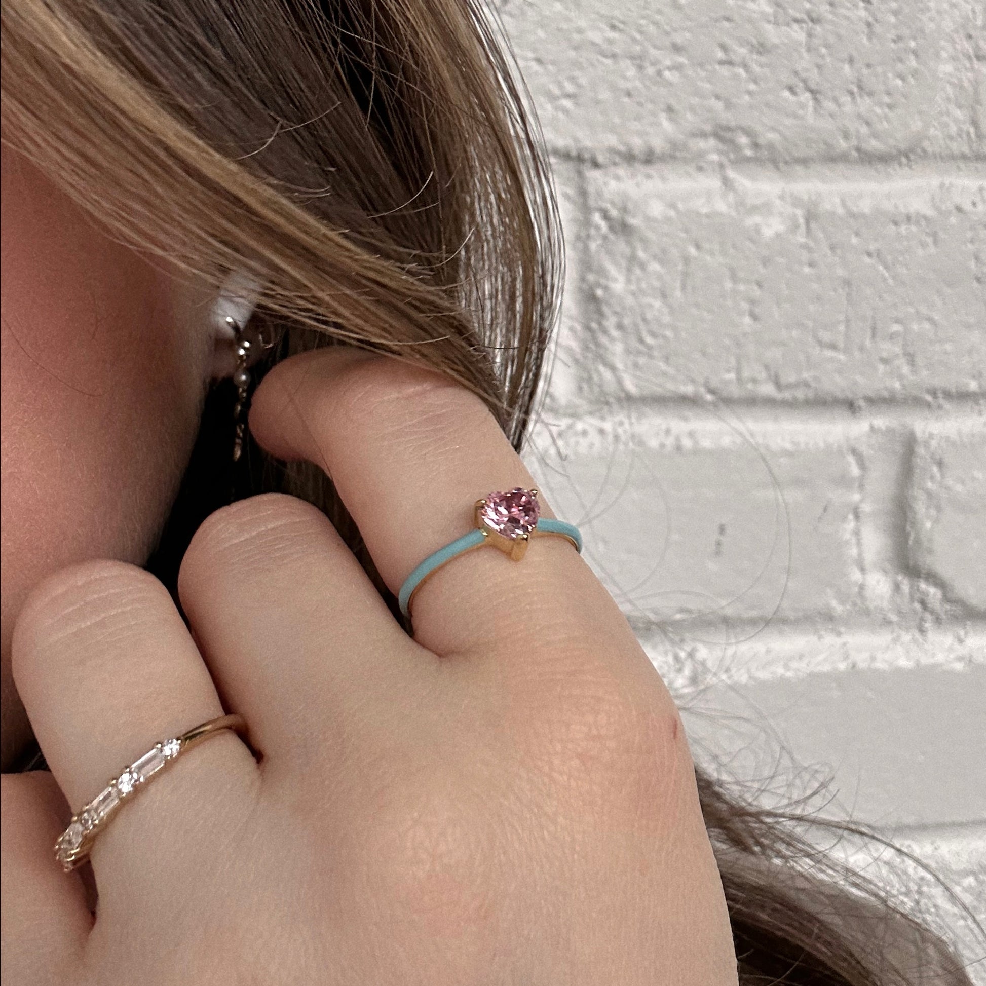 A close-up of a womans hand near her face features the Little Love Ring with a pink heart-shaped stone on a light blue band, paired with a 14k yellow gold plated thin band with small clear stones. She stands against a white brick wall.