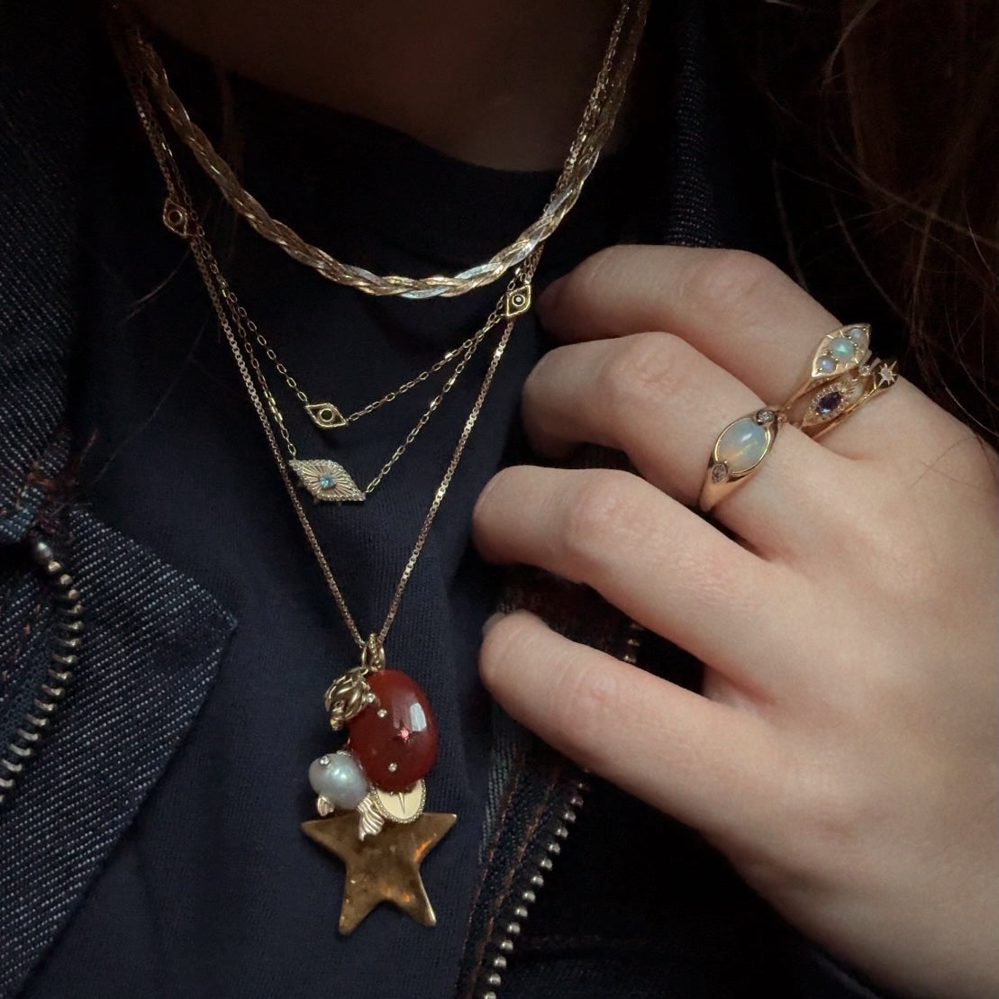 A hand with gold rings featuring opal-like stones rests on a chest layered in gold necklaces, including the Solid Gold Zodiac Constellation Crystal Charm with a star and red stone pendant, over a dark shirt and denim jacket.