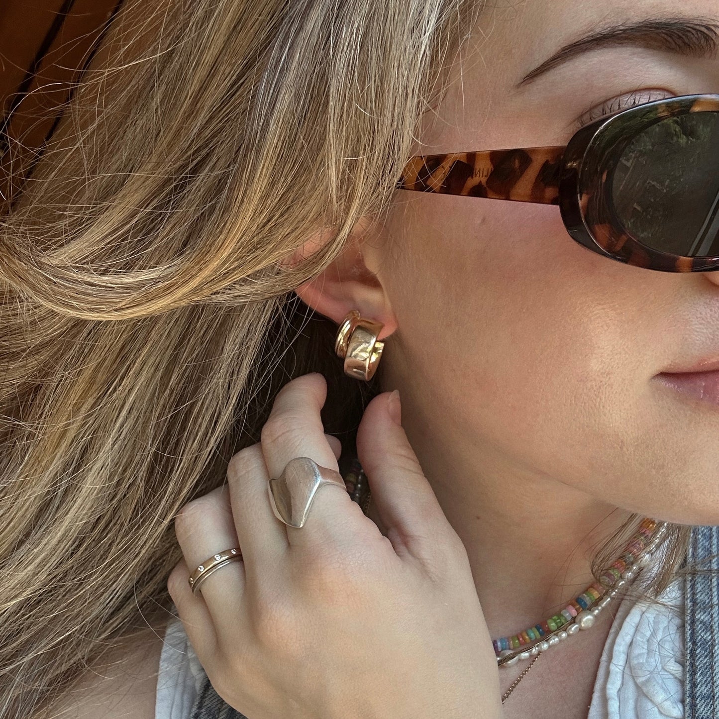 A blonde woman wears tortoiseshell sunglasses, Solid Gold Twisted Hollow Hoops, a silver heart-shaped ring, a thin gold ring, and a colorful beaded necklace. She faces sideways and touches her earring.