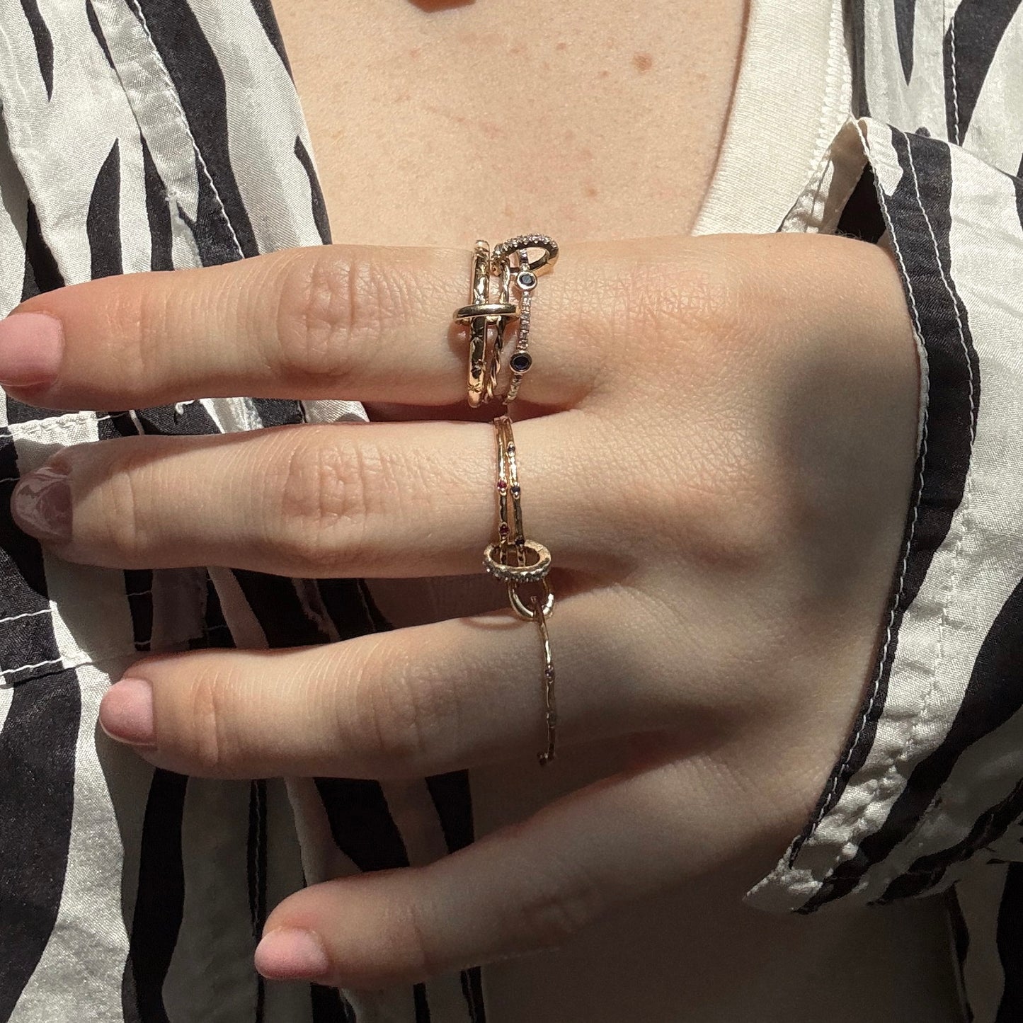 A hand with light skin wears the Solid Gold Ring Clip, a customizable ring stack of unique gold rings joined by a chain on the middle and ring fingers, paired with a black and white patterned shirt.